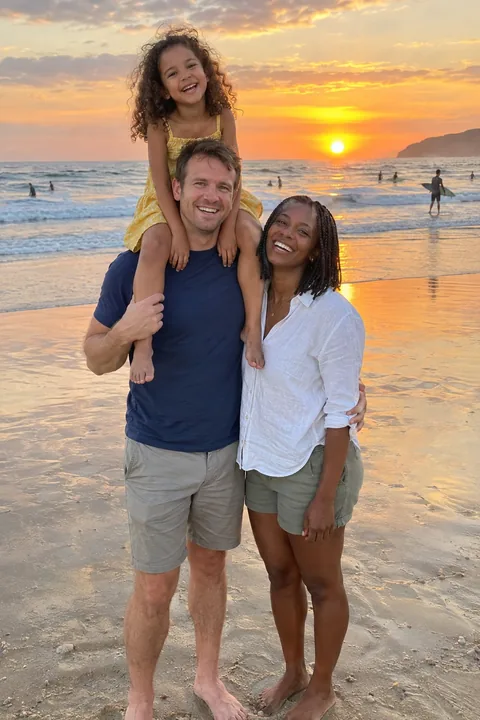 A holiday photo of a family on a beach at sunset, the daughter on her dad's shoulders